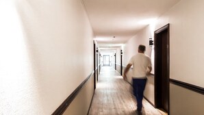 A hotel hallway with wooden flooring, a person walking, and a wall-mounted light fixture. - Hotelli Havu (Uusikaupunki)