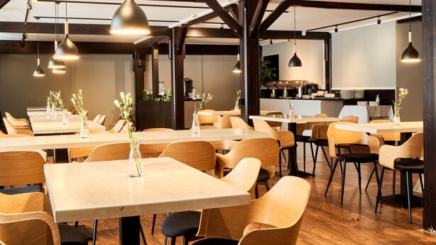 A restaurant with wooden tables and chairs, pendant lighting, and a visible kitchen area. - Hotelli Havu (Uusikaupunki)