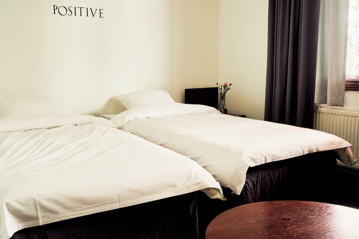 Ein Hotelzimmer mit zwei Betten, einem Nachttisch mit einer Vase, einem Fenster mit Vorhängen und einem Heizkörper.