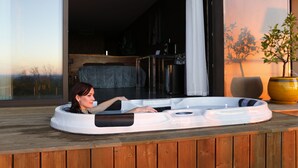 Una persona relajándose en un jacuzzi en una terraza de madera, con vista a un interior moderno y una planta en maceta.