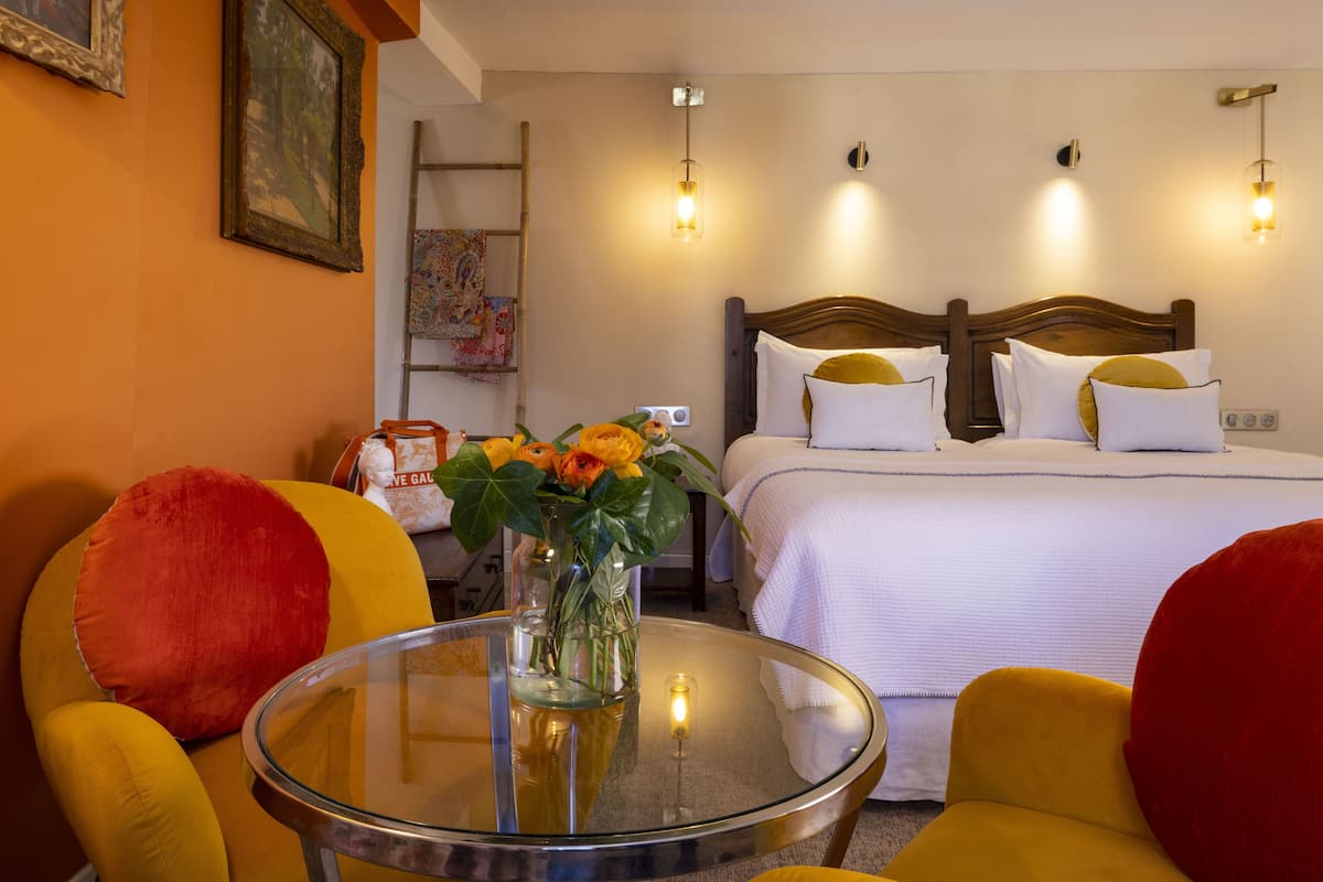A hotel room with a bed, two chairs, a small table with a vase of flowers, and a painting on the wall.