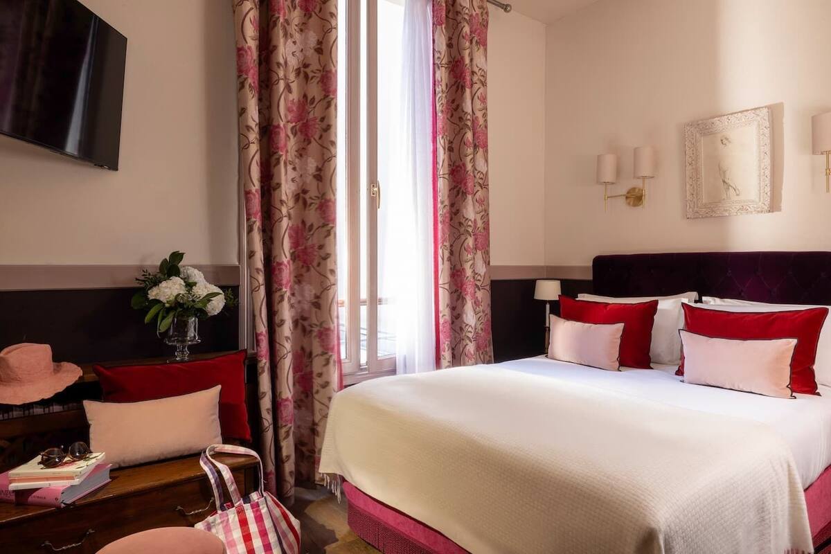 A hotel room with a large bed, a desk with a chair, a TV mounted on the wall, and a window with floral curtains.