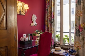 A room with a red wall, a bust sculpture, a desk with a coffee maker, a vase with flowers, and a window with floral curtains.