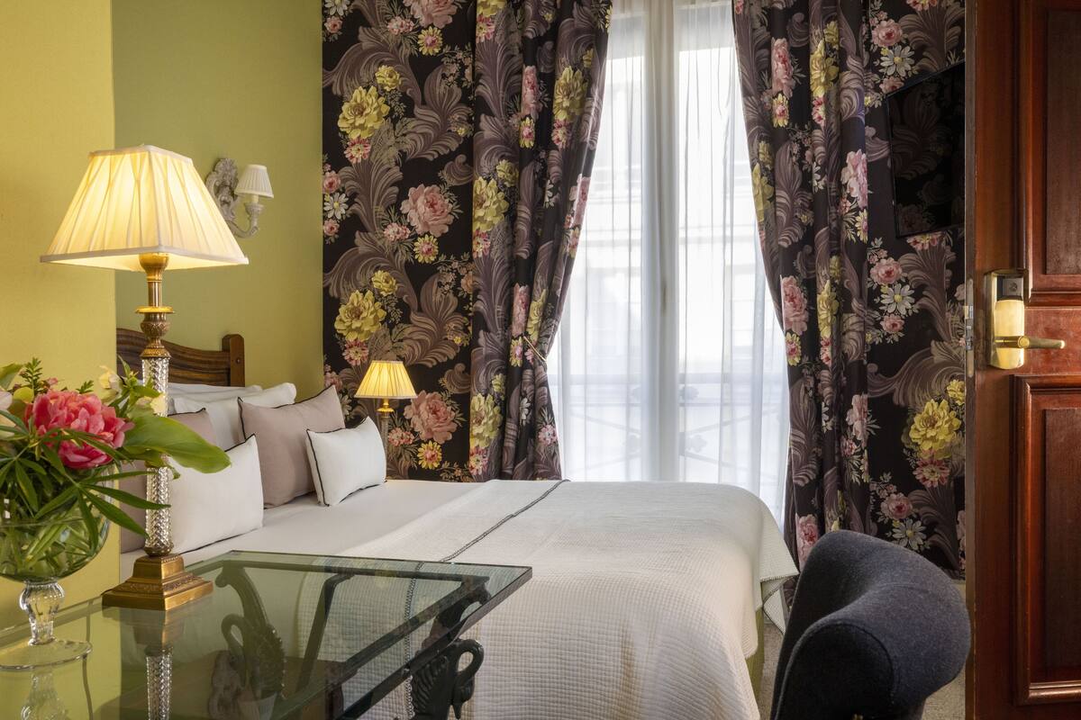 A hotel room with a bed, two bedside lamps, a glass table with a vase of flowers, and floral curtains.