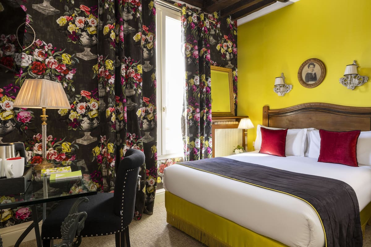 A hotel room with a bed, a desk, and a chair. There is a window with curtains, a lamp, and a picture on the wall.