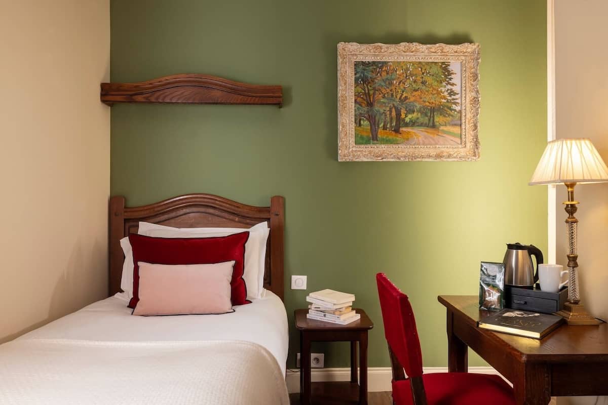A hotel room with a wooden bed, a red chair, a desk with a lamp, a painting on the wall, and a coffee maker.