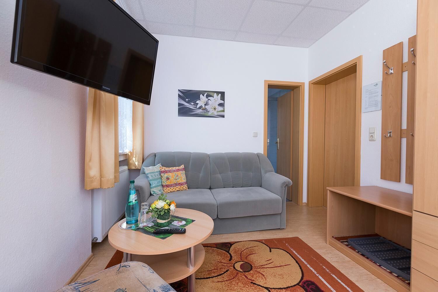 A small living room with a grey sofa, a round coffee table, and a TV mounted on the wall. There is a wooden cabinet and a door leading to another room.