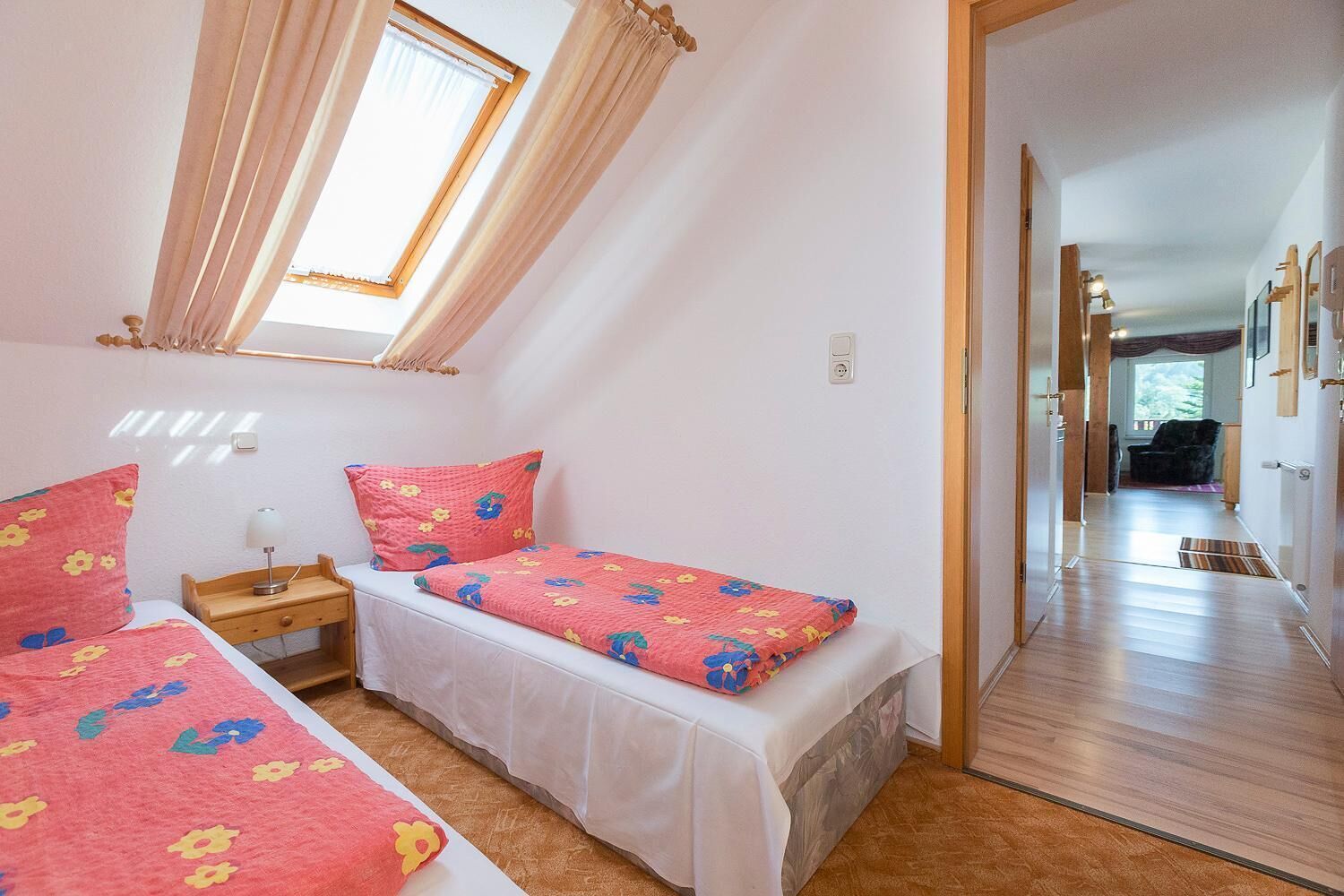 A bedroom with two beds, a window with curtains, a bedside table with a lamp, and a hallway leading to another room.