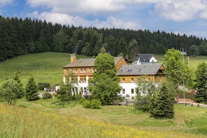 Uma casa grande, com vários andares e telhado inclinado, cercada por uma paisagem verde exuberante e uma floresta densa ao fundo.