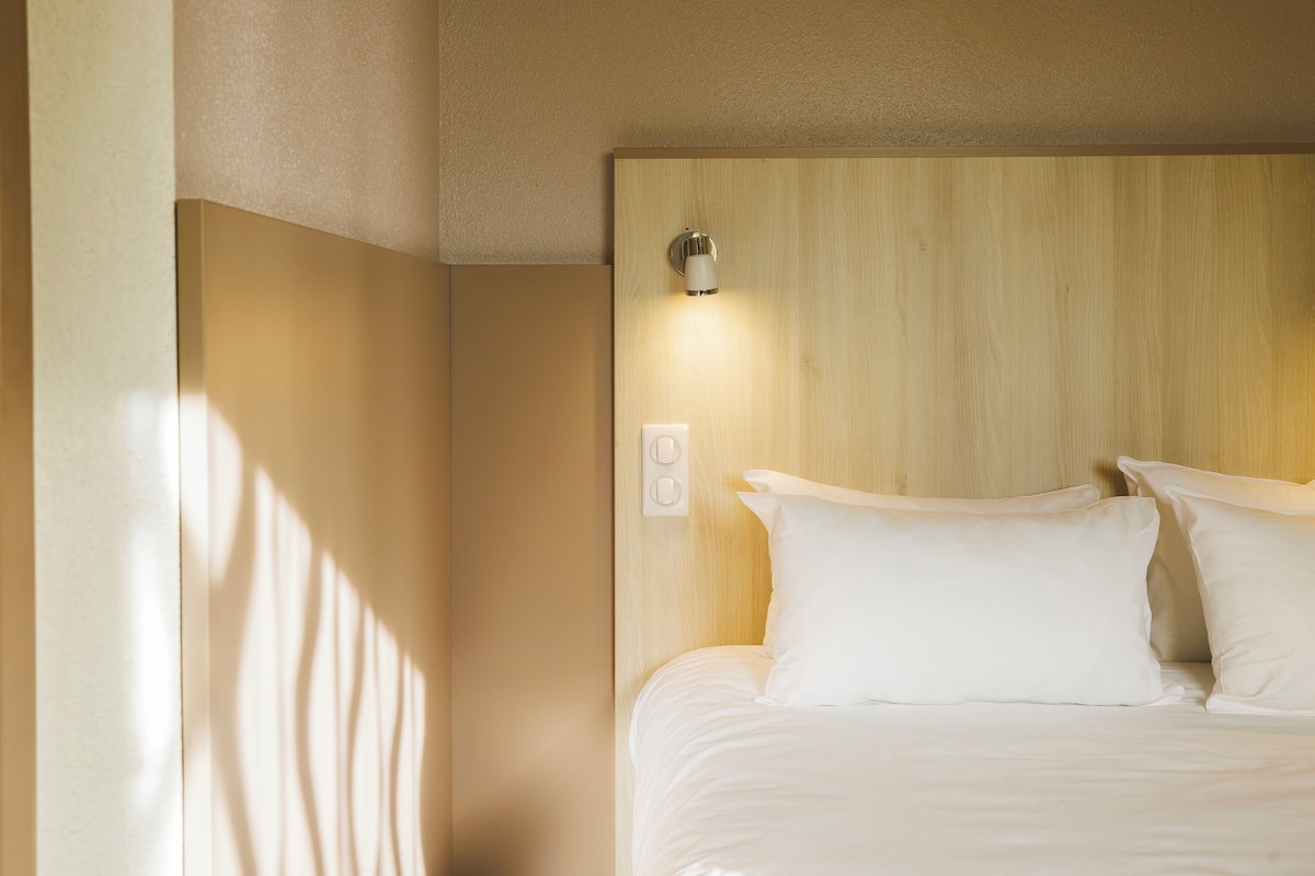A neatly made bed with white linens and pillows, a wooden headboard, and a wall-mounted light fixture.