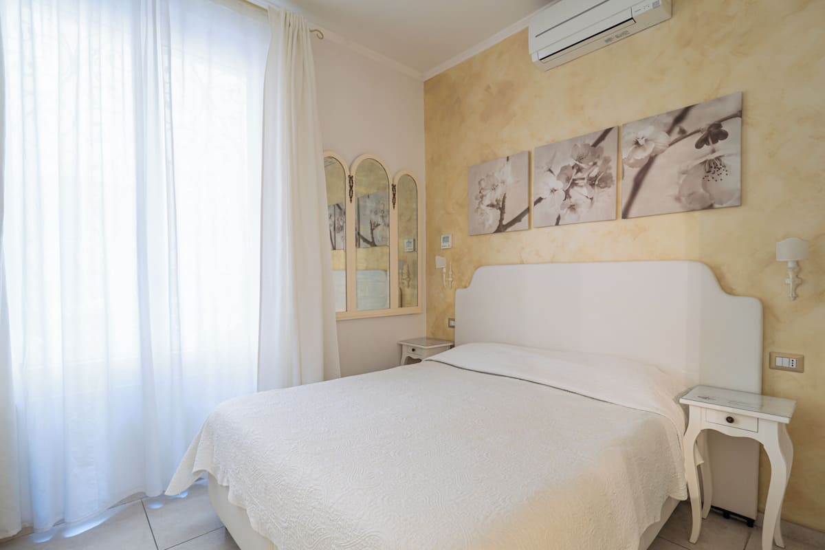 Une chambre avec un grand lit, une table de chevet, une commode, un climatiseur fixé au mur et une fenêtre avec des rideaux légers.
