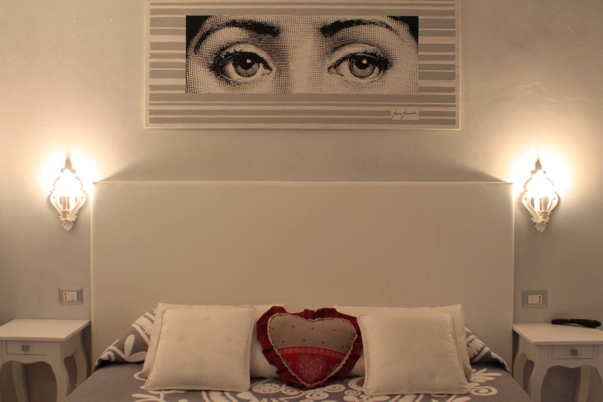 Une chambre à coucher comprenant un lit, deux tables de chevet, un luminaire fixé au mur et un tableau encadré au-dessus du lit.