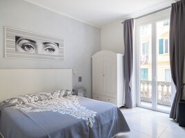 Un dormitorio con cama, un armario blanco, una puerta y vista a un edificio por la ventana.