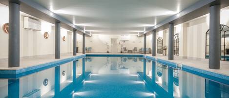 A large indoor swimming pool with clear blue water, surrounded by columns and a tiled floor.