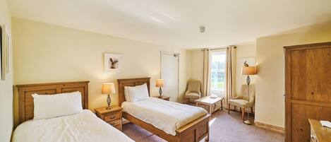 A hotel room with two beds, a wooden wardrobe, a desk, two chairs, and a lamp.