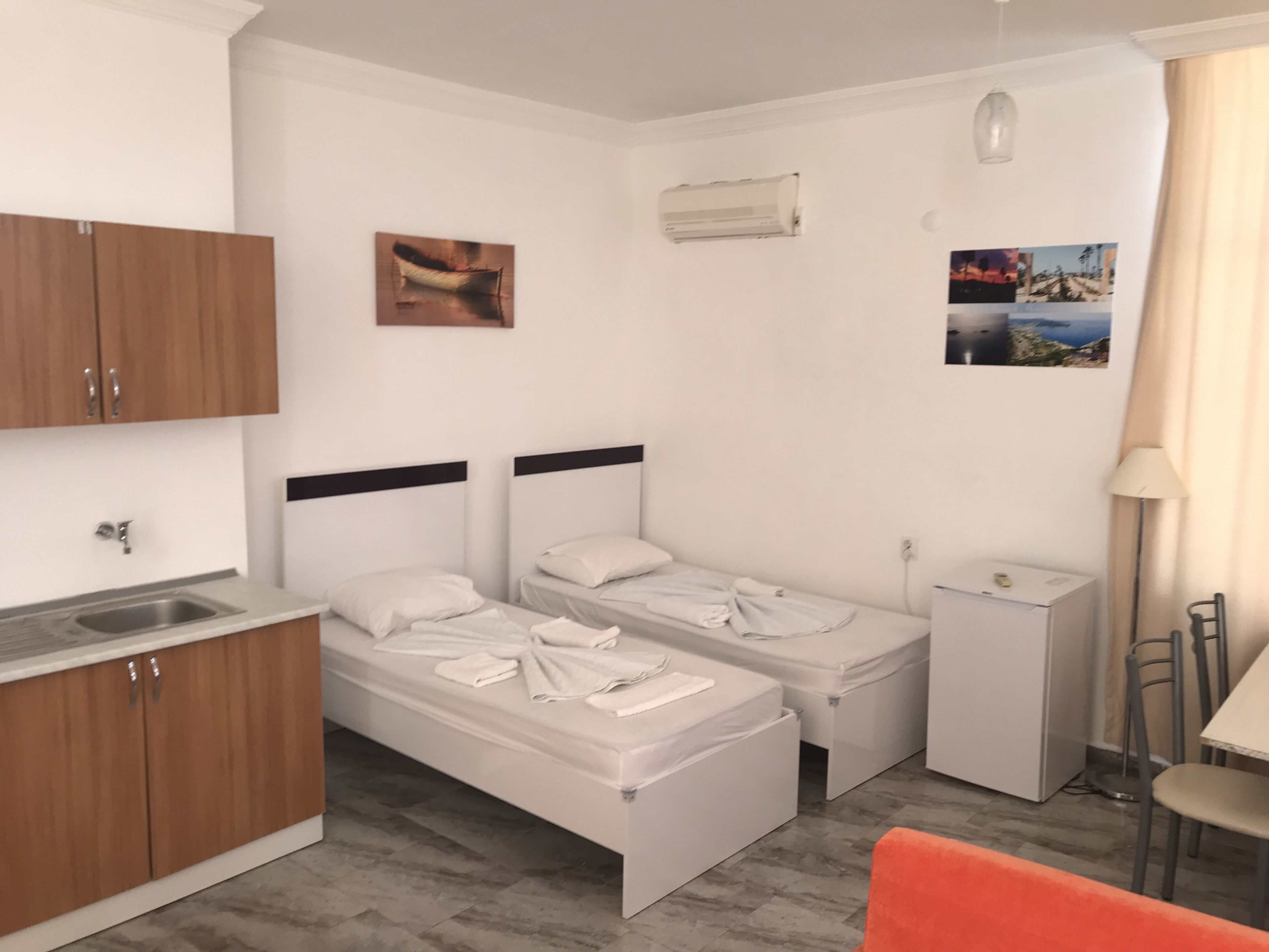 A compact hotel room with two single beds, a small kitchenette, a desk, and a chair.