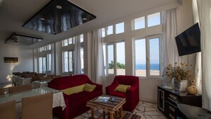 A spacious living area with a red sofa, two armchairs, a coffee table, and a TV on a stand. Large windows offer a view of the sea. - Pinar Motel 2022-33-0198 (Aydincik)