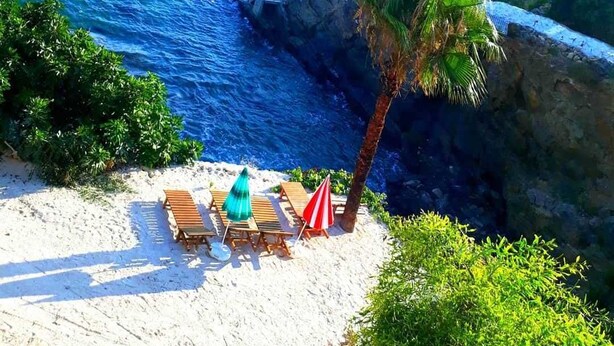 A beach with wooden lounge chairs and a palm tree. - Pinar Motel 2022-33-0198 (Aydincik)