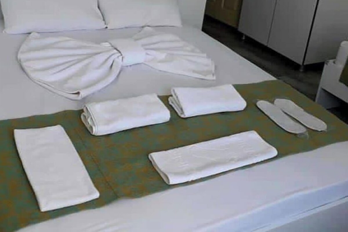 A neatly made bed with white linens, a folded towel, and a pair of slippers on a bed with a green bedspread.