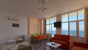 A modern hotel room with a large window offering a sea view, a flat-screen TV mounted on the wall, a dining area with a table and chairs, and a seating area with a sofa and armchairs. - Pinar Motel 2022-33-0198 (Aydincik)