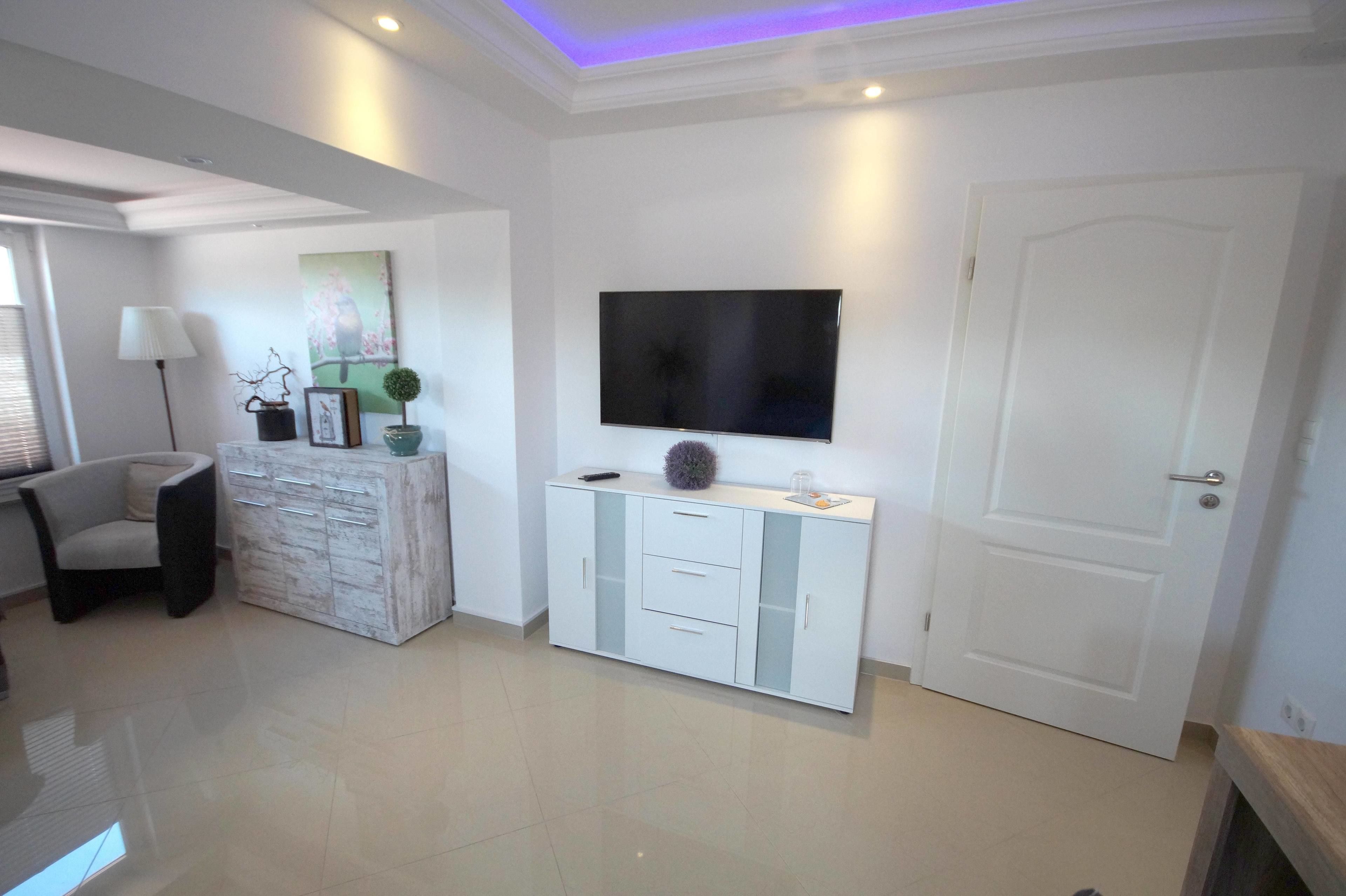 A modern living room with a flat-screen TV mounted on the wall, a white cabinet with drawers, a grey armchair, and a door.