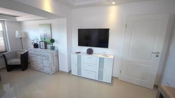 A modern living room with a flat-screen TV mounted on the wall, a white cabinet with drawers, a grey armchair, and a door.