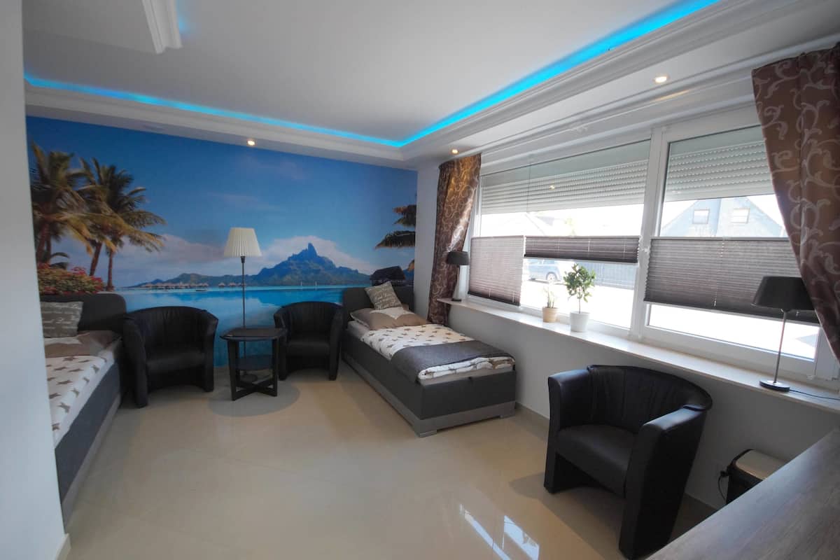 Um quarto com uma cama, duas poltronas, uma mesinha, uma janela com persianas, um abajur e um mural com uma paisagem de praia.