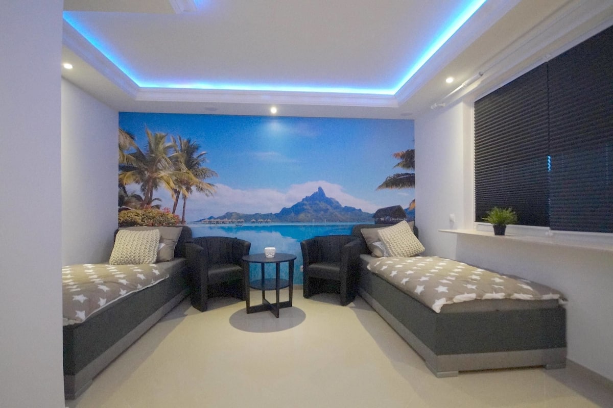 Quarto com um mural de uma praia tropical, dois sofás, uma mesa pequena e uma janela com persianas.