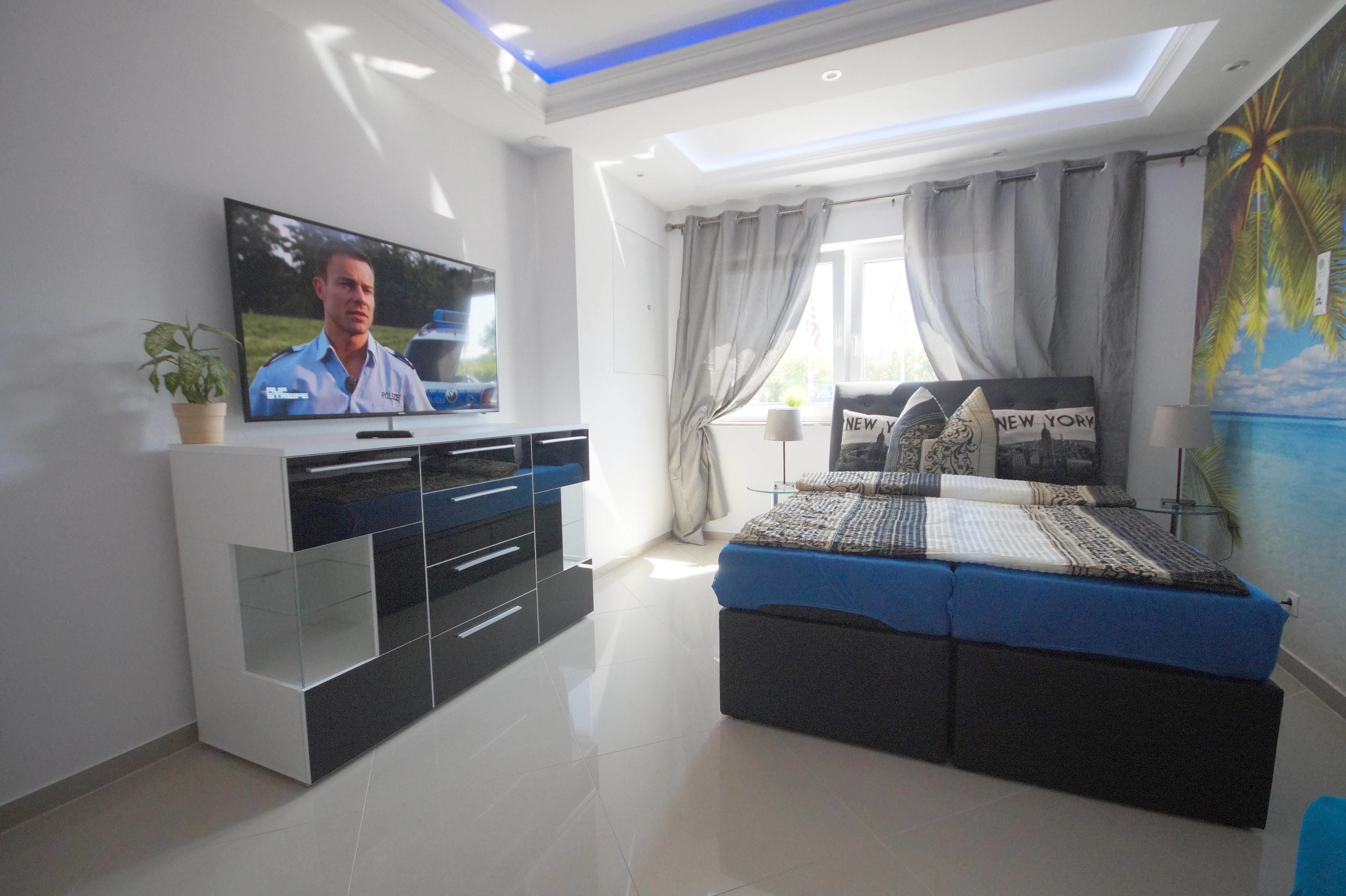 Un dormitorio moderno con un televisor de pantalla plana grande, una cama con colcha azul y blanca, una mesita de noche con lámpara y una ventana con cortinas.