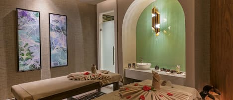 A spa room with two massage tables, a green wall, a sink, and framed artwork.