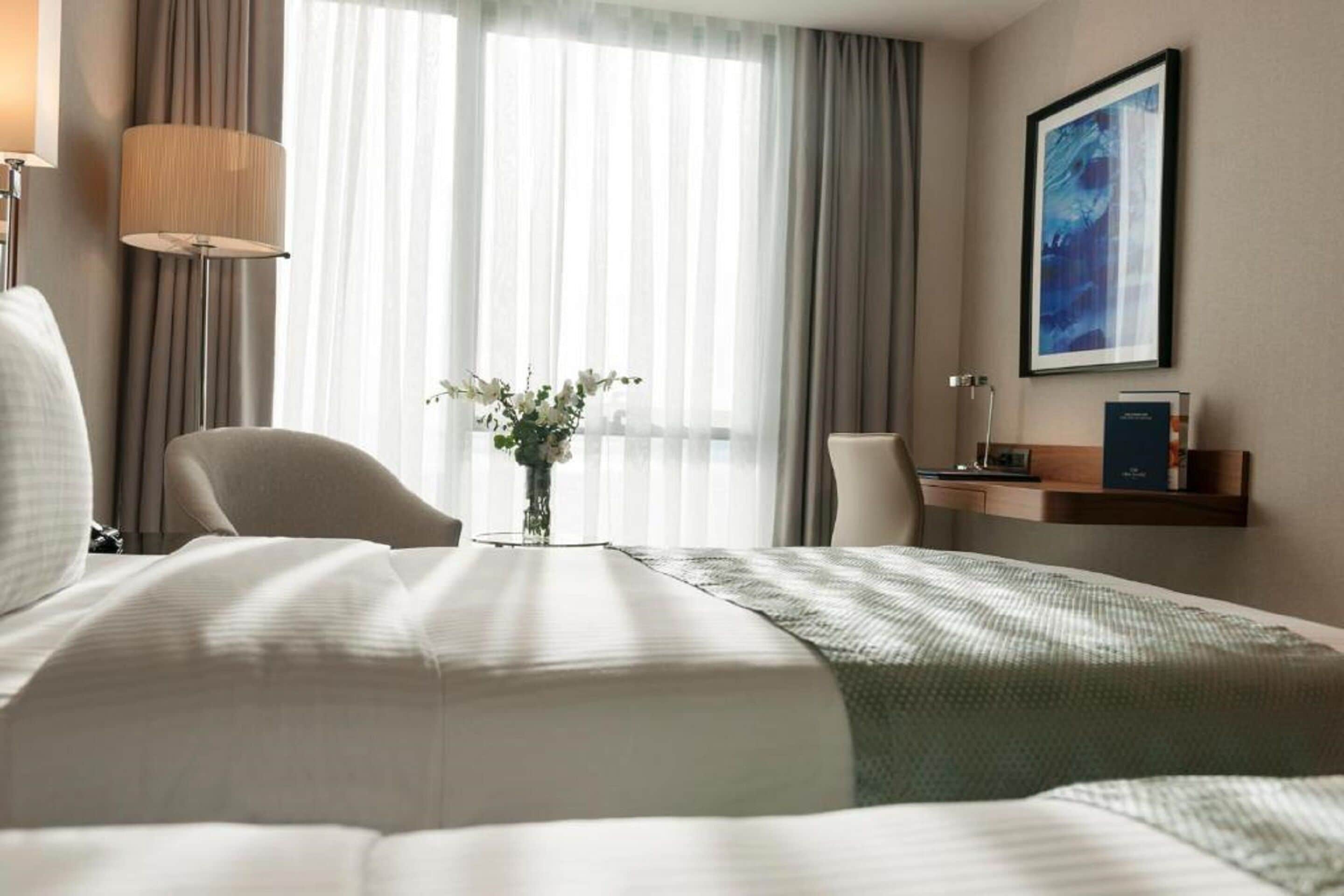 A hotel room with a large bed, a chair, a desk, a lamp, a vase with flowers, and a framed picture on the wall.