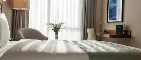 Habitación de hotel con una cama grande, una silla, un escritorio, una lámpara, un florero con flores y un cuadro en la pared.