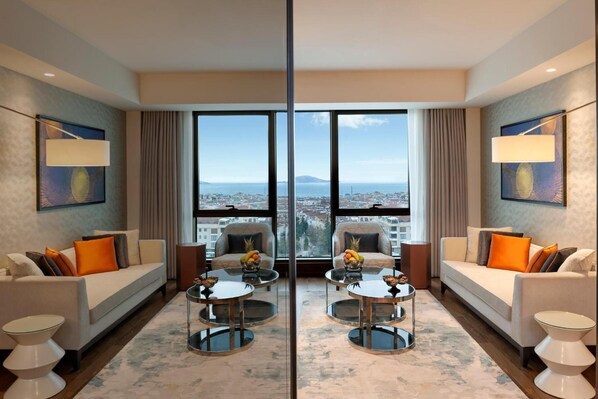 Habitación de hotel moderna con un ventanal grande que ofrece vistas a la ciudad, dos sofás con cojines anaranjados y una mesa de centro con fruta.