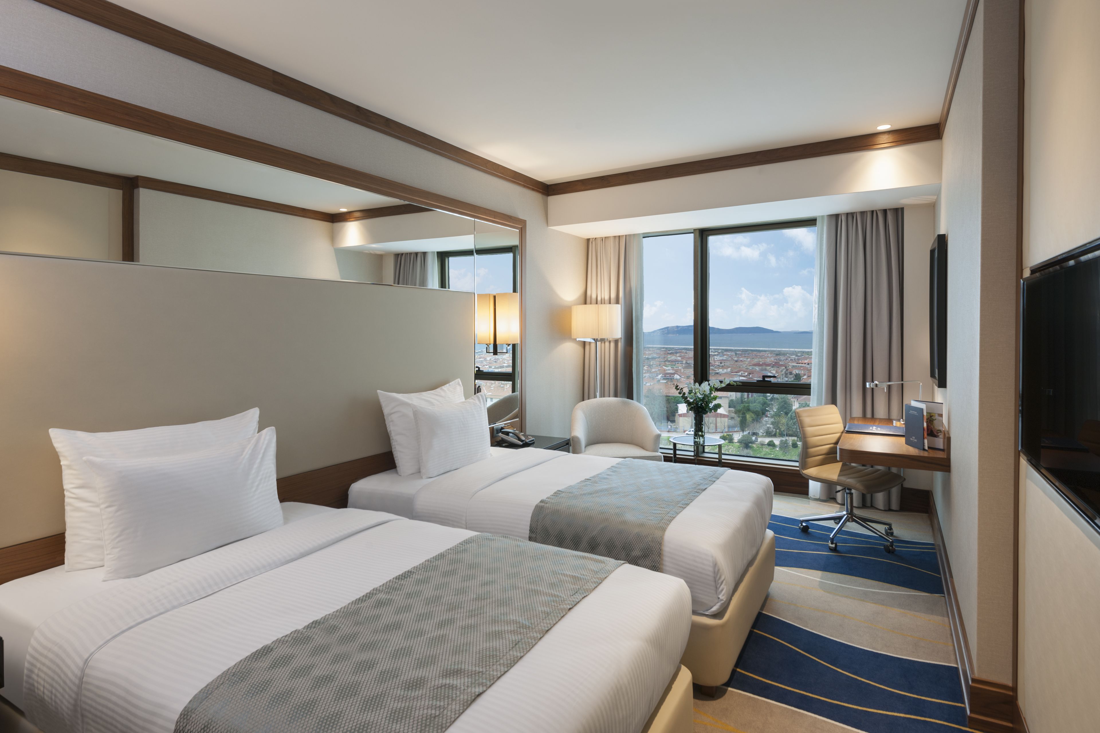 Een hotelkamer met twee bedden, een bureau met een computer, een stoel, een televisie en een groot raam met uitzicht op de stad.
