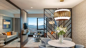 A modern living room with a large window, a dining area with blue chairs, and a staircase with a decorative light fixture. - Elite World Grand Istanbul Kucukyali  (Istanbul)