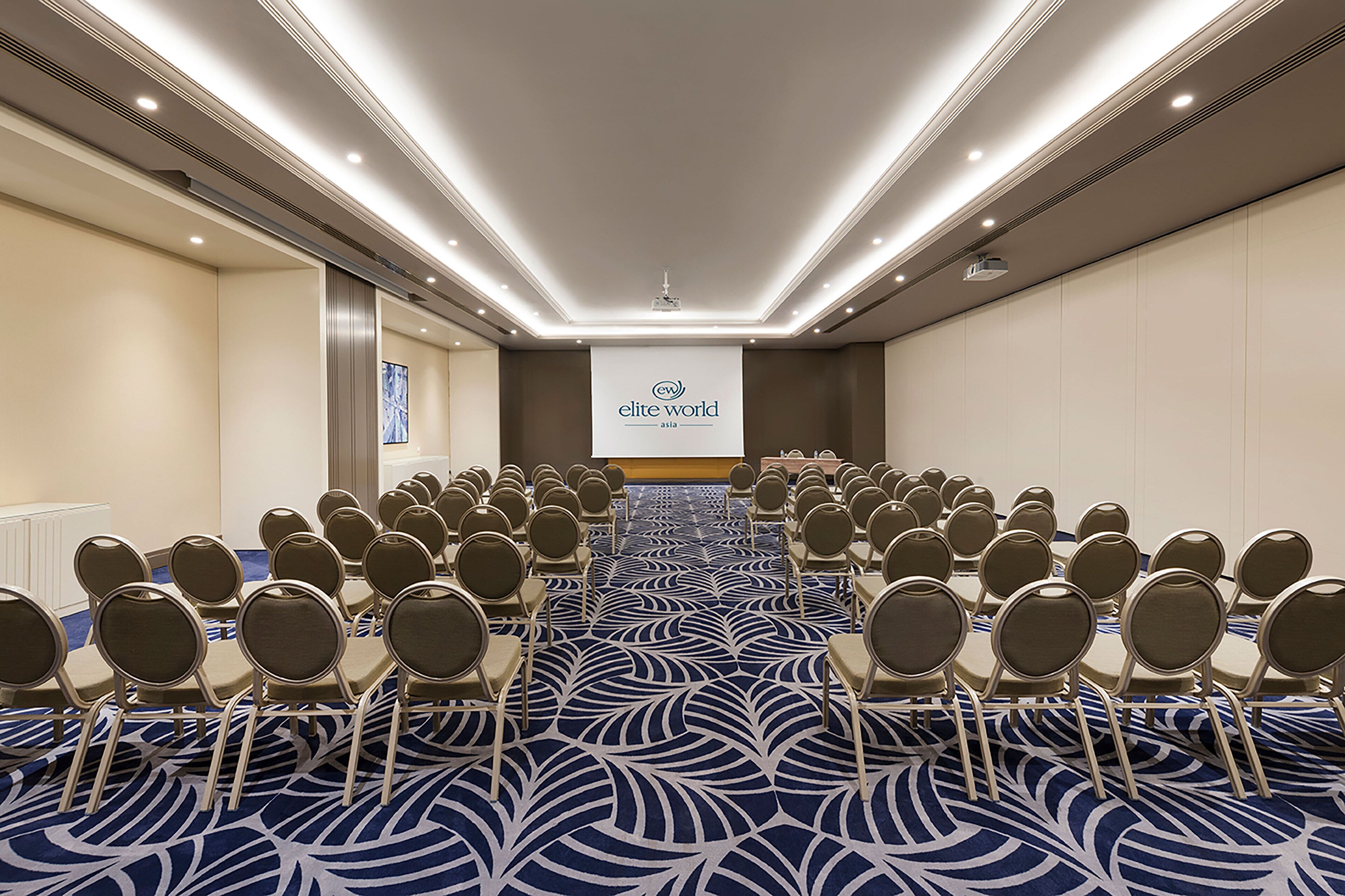 A conference room with rows of chairs facing a presentation screen, a patterned carpet, and a modern ceiling design.