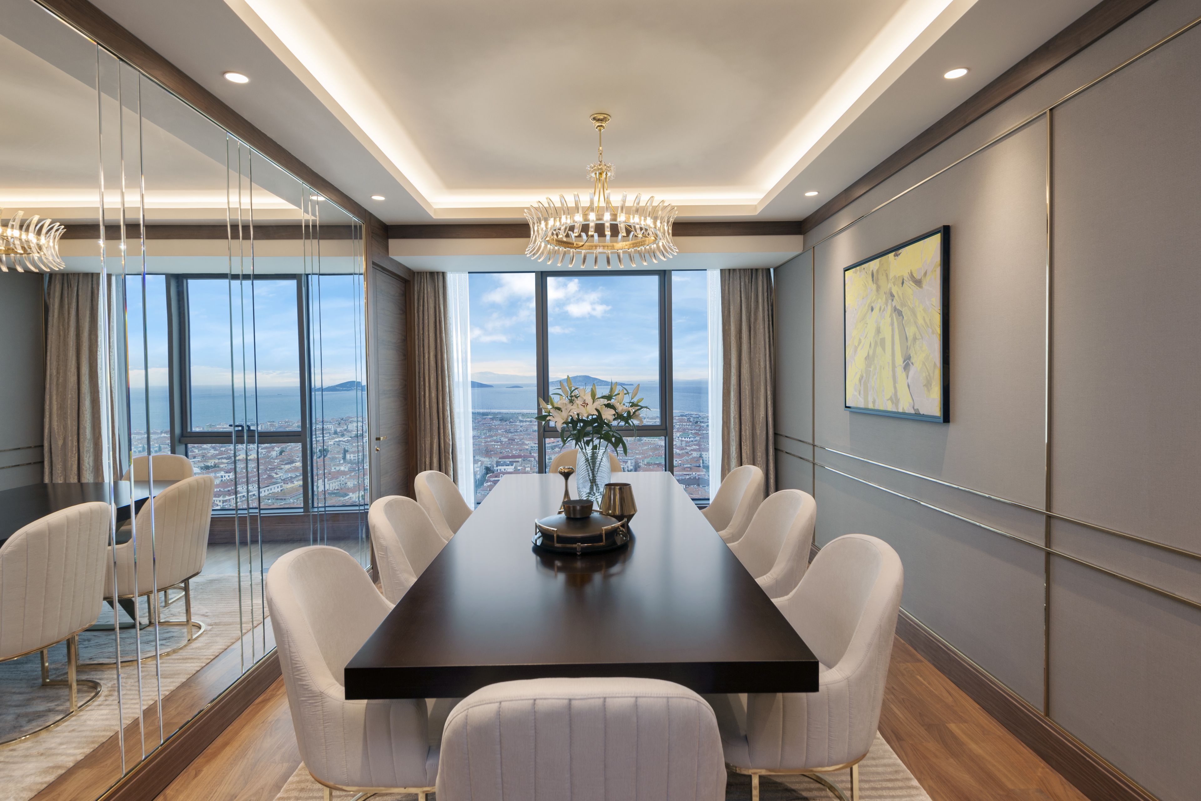 A conference room with a long table, white chairs, a chandelier, and a large window offering a city view.