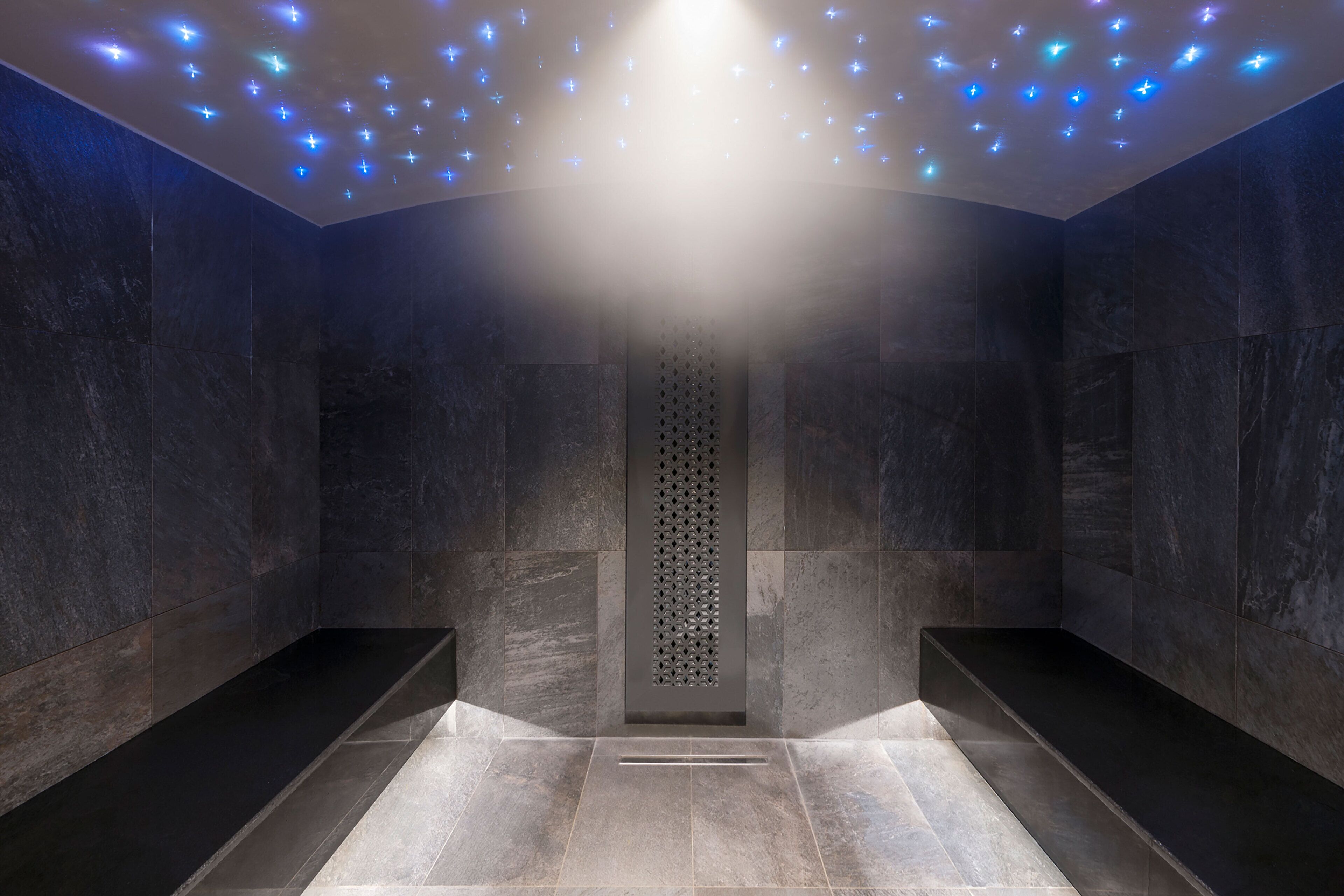 Un baño moderno con paredes de piedra oscura, una columna central y un techo con luces azules.