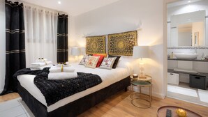 Ein modernes Schlafzimmer mit einem großen Bett, zwei Nachttischlampen, einem Nachttisch mit Kerze und Blick ins Badezimmer durch eine offene Tür.