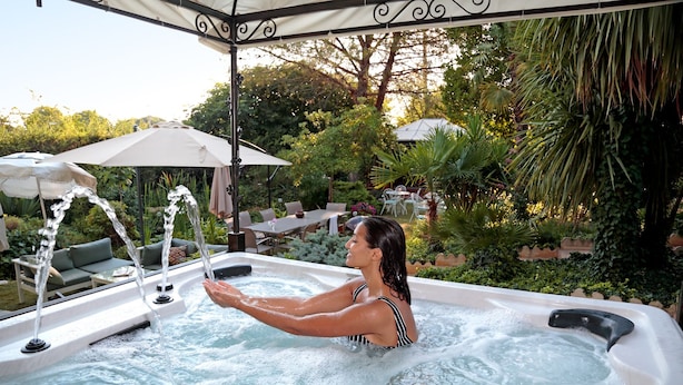 Ein Whirlpool mit einer Person darin, umgeben von Gartenmöbeln und viel Grün.