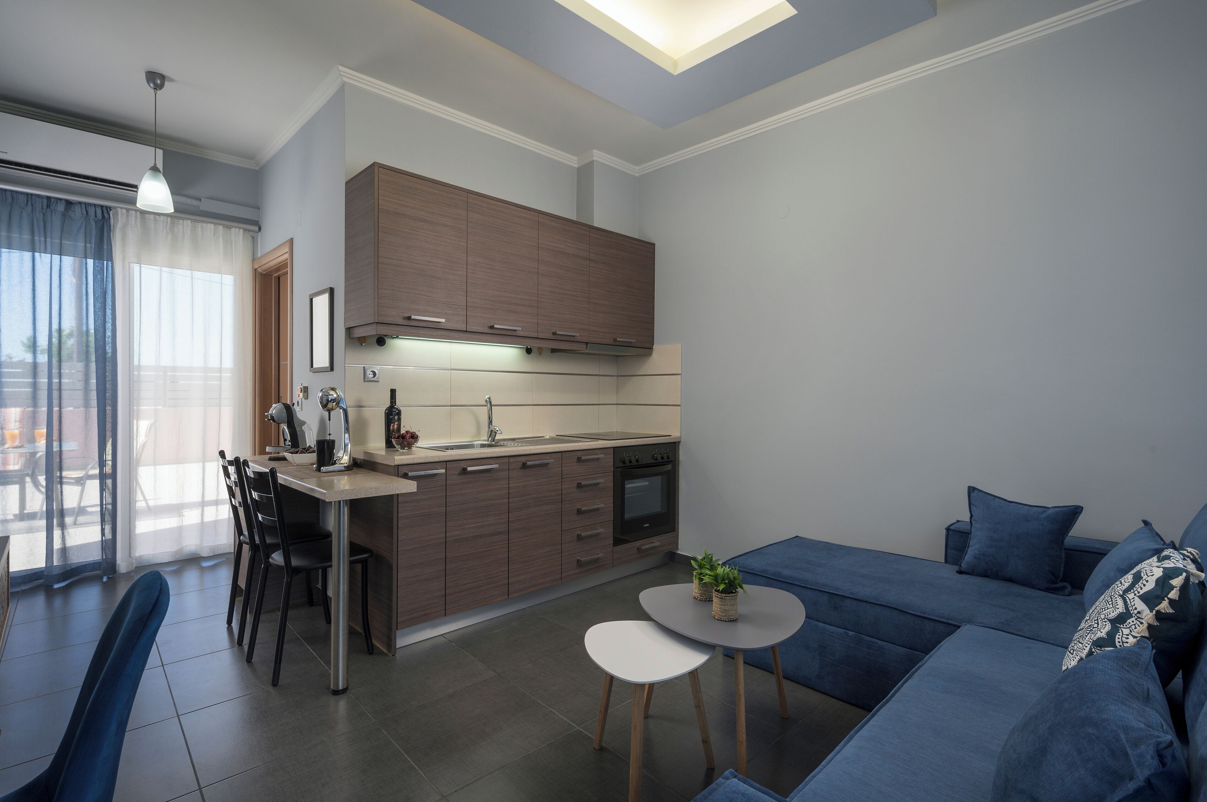 A modern kitchen with wooden cabinets, a dining area with a table and chairs, a blue sofa, and a small round table with two potted plants.