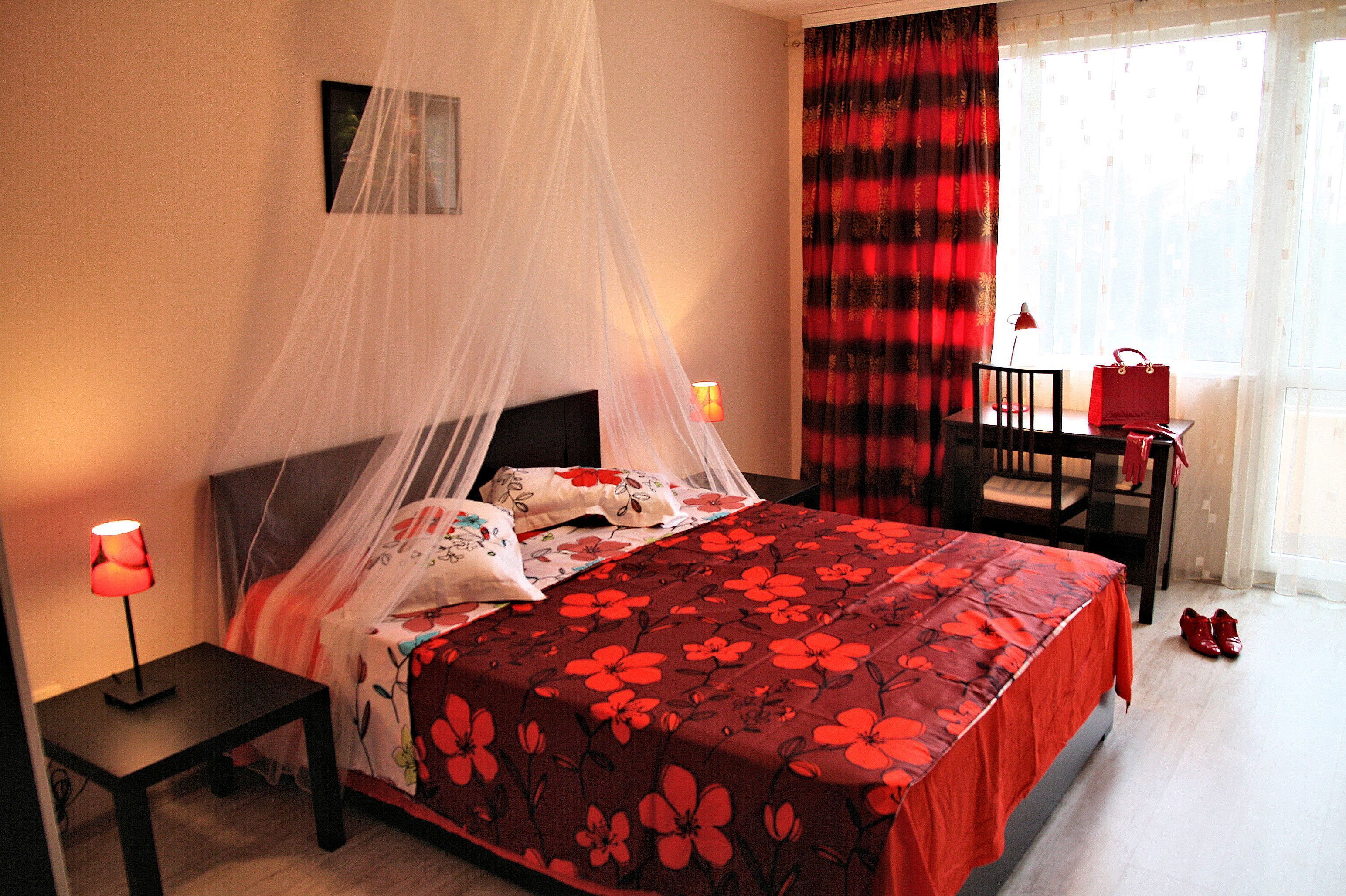 A neatly made bed with a floral bedspread, a bedside table with a lamp, a chair, and a window with curtains.