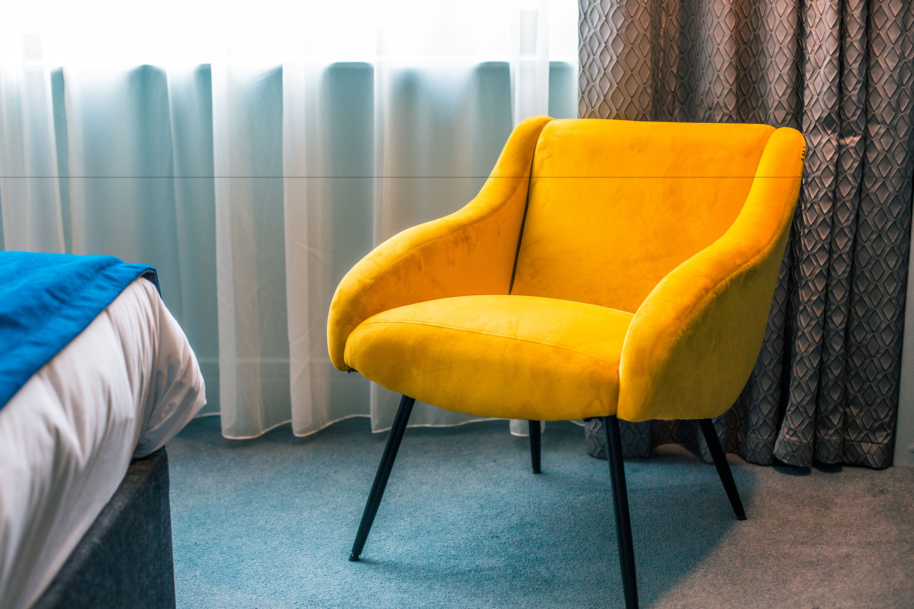 A room with a yellow armchair, a bed with a blue blanket, and a window with sheer curtains.