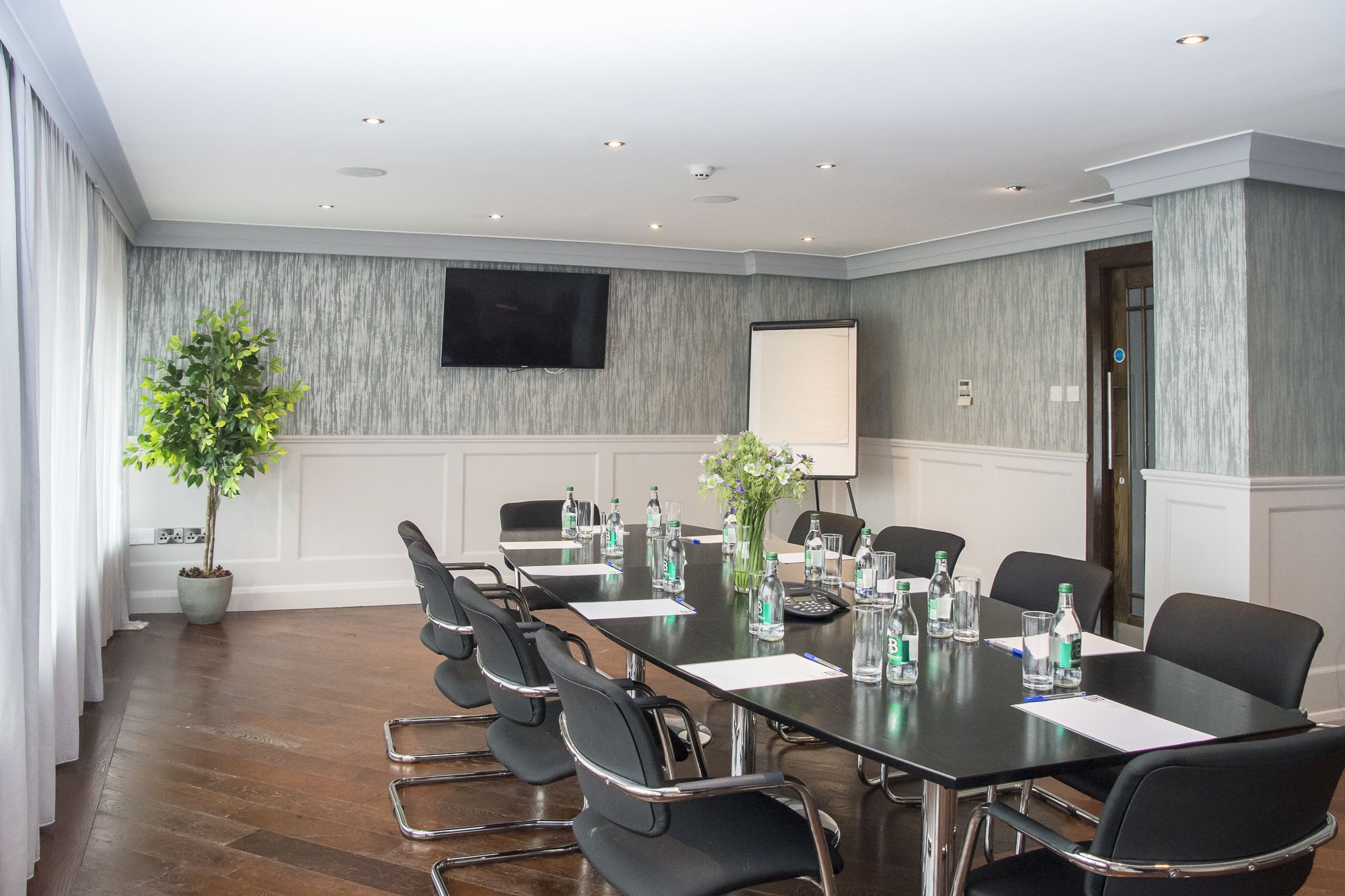 A conference room with a long table, chairs, a projector screen, a potted plant, and a television on the wall.