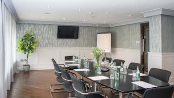 A conference room with a long table, chairs, a projector screen, a potted plant, and a television on the wall.