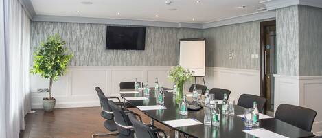 Un salón de conferencias con una larga mesa, sillas, una pantalla de proyector, una planta en maceta y un televisor en la pared.