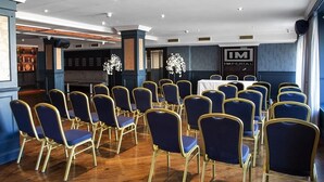 Uma sala de conferências com fileiras de cadeiras azuis e uma mesa, pisos de madeira, e uma placa com a inscrição 'IMPERIAL'.