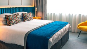 Quarto de hotel com uma cama grande, mesa de cabeceira com abajur, uma cadeira amarela e vista para o exterior através de cortinas leves.