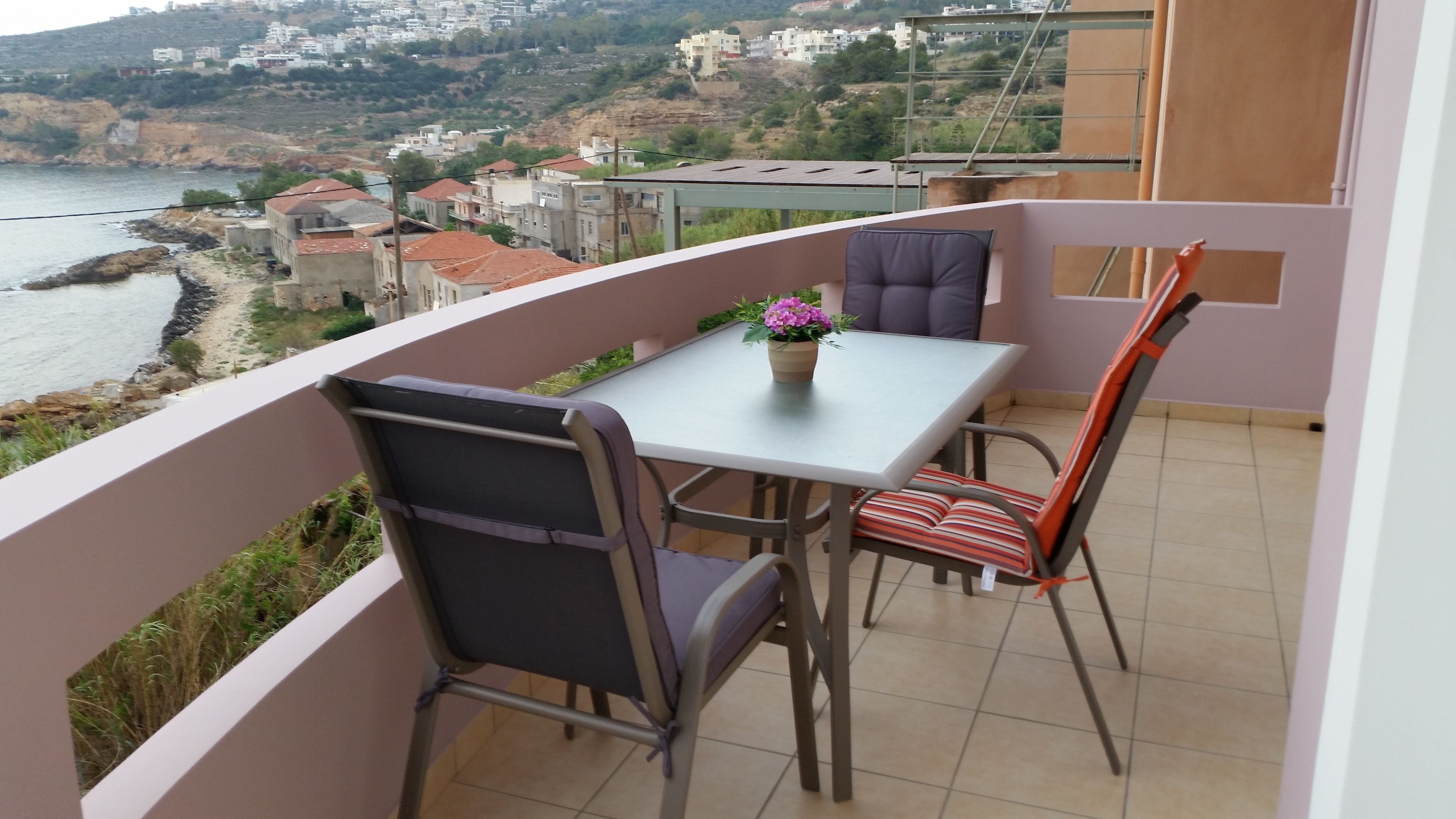 A balcony with a table and chairs, overlooking a coastal town with buildings and a body of water.