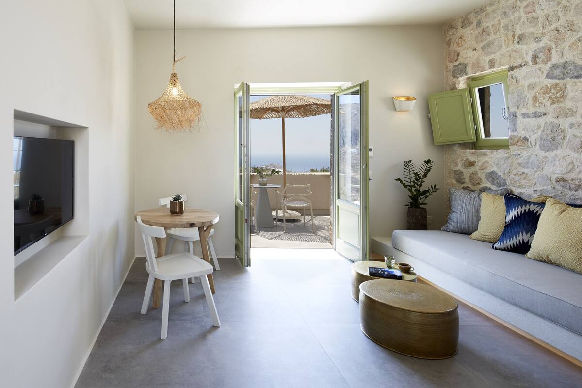 A modern living room with a stone wall, a sofa with patterned cushions, a dining area with a table and chairs, and a view of the sea through an open door.